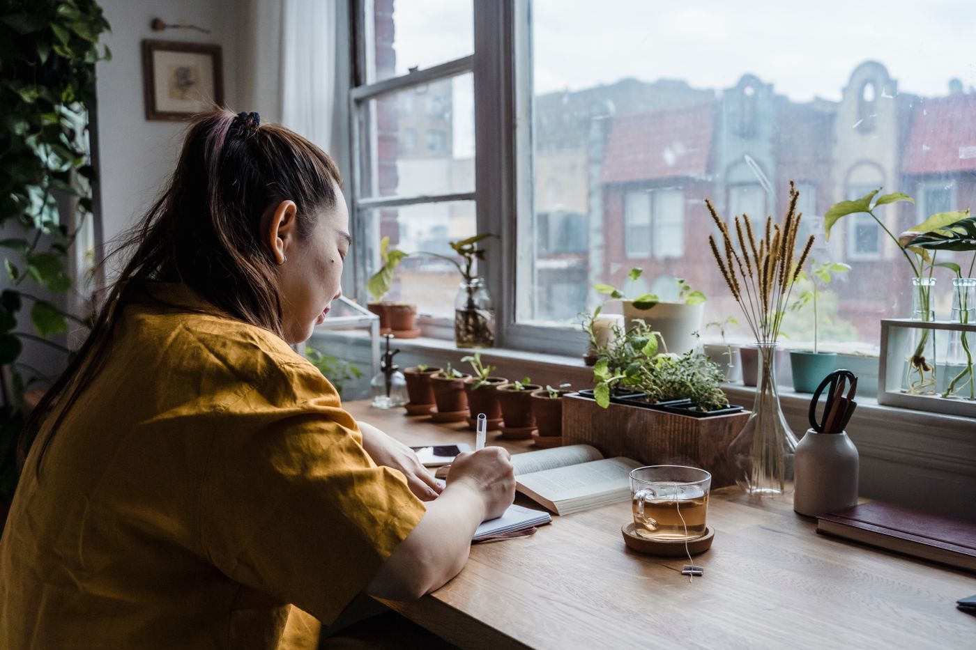Una mujer escribe en un cuaderno junto a una ventana y plantas